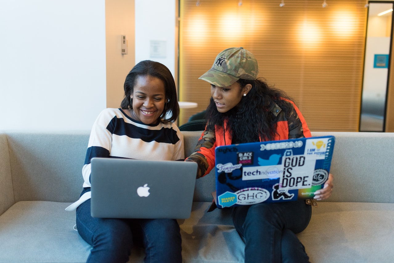 Two black women sitting on a couch, each on their laptop, talking to each other.