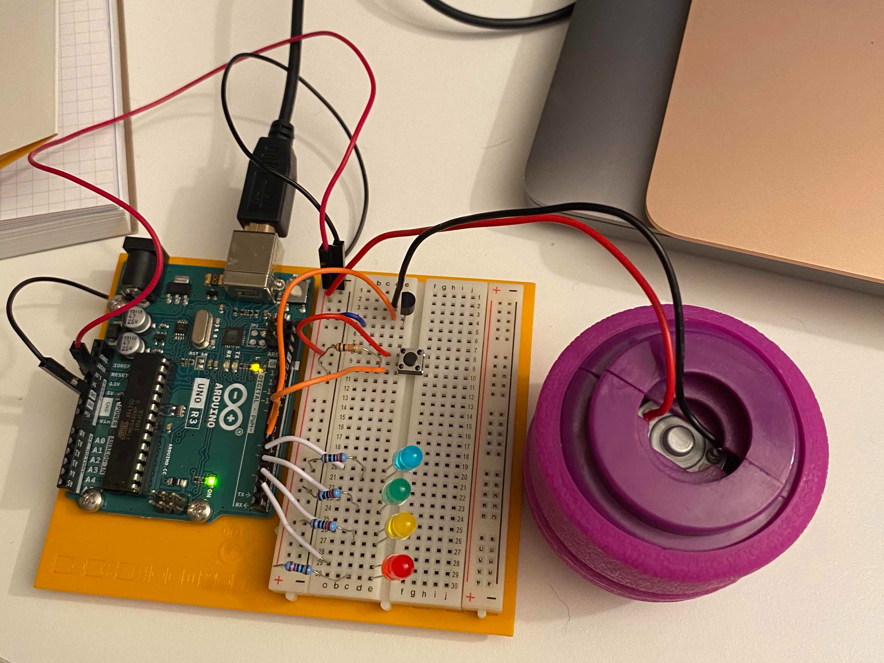 photo of an arduino and breadboard circuit with 4 colored LEDs, a button, and a purple circular object. There is a laptop in the upper left corner